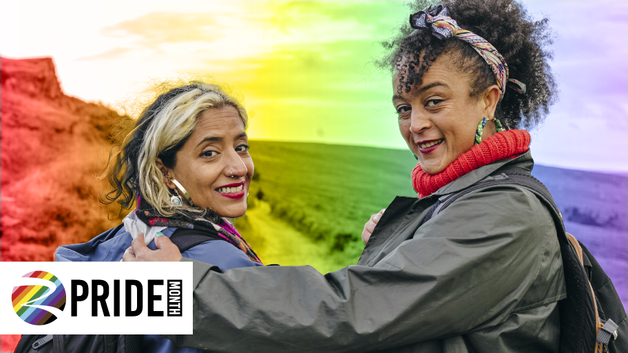 Two people smiling outdoors in front of a rainbow-colored landscape, celebrating Pride Month